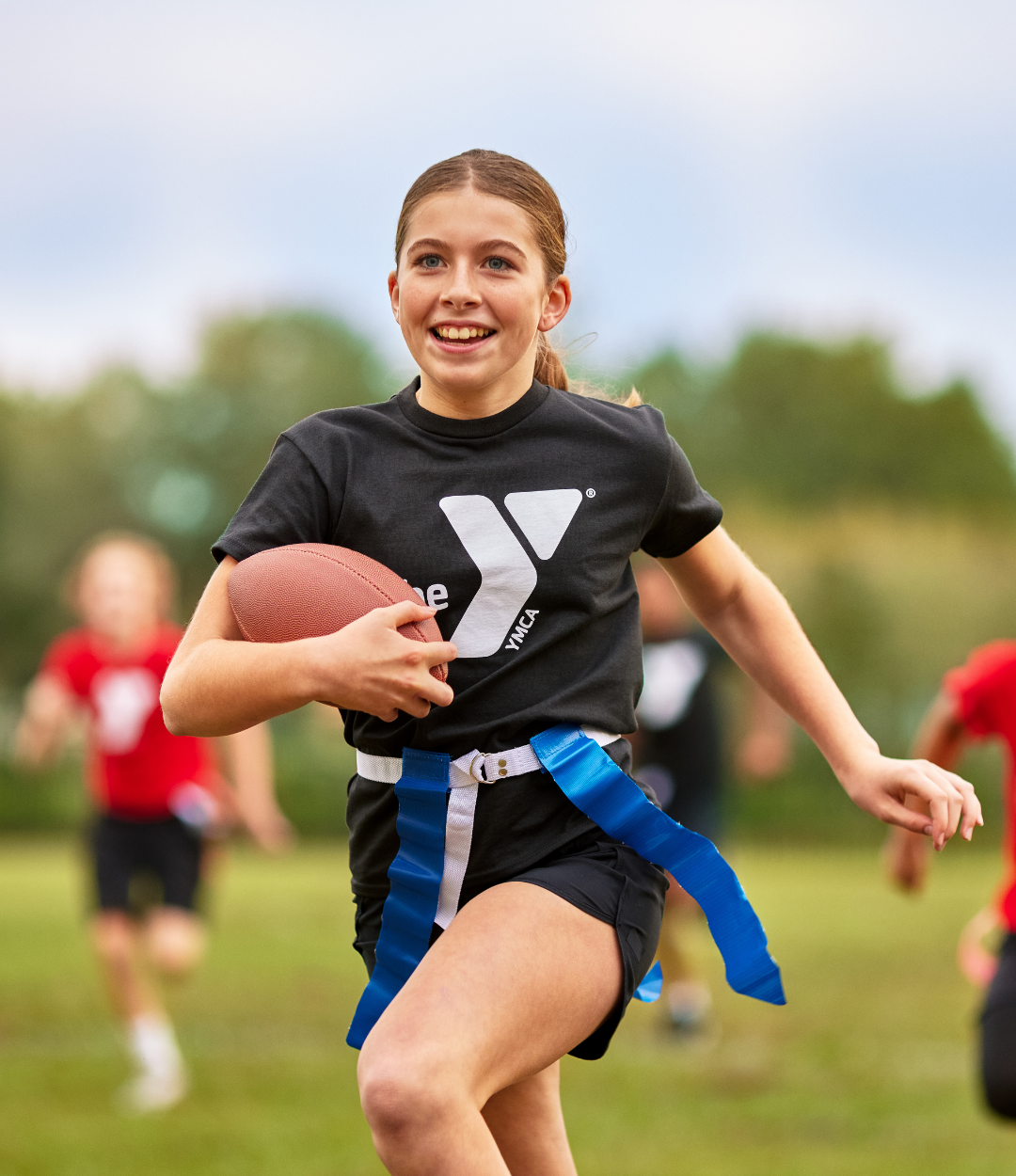 Four teenagers run down the flag football field during an outdoor game with one at the front carrying the football.